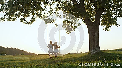 LENS FLARE SILHOUETTE: Unrecognizable couple swaying on swing at golden twilight Stock Photo