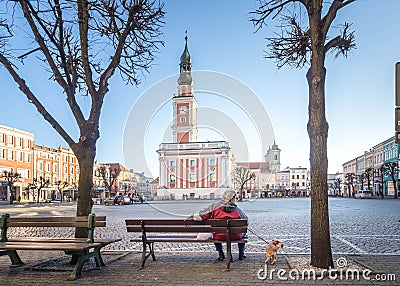 LESZNO, POLAND - FEBRUARY 16, 2019. An older man with a dog on a lead sitting on a bench in front of the Leszno Town Hall. Editorial Stock Photo