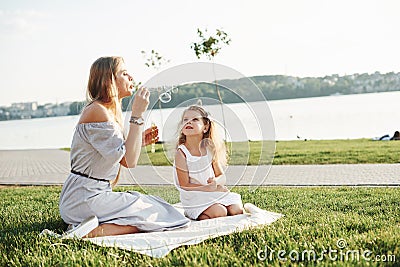 Let the launch of bubbles has come. Photo of young mother and her daughter having good time on the green grass with lake Stock Photo