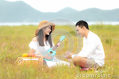 Lifestyle couple picnic sunny time. Asian young couple having fun and relax playing guitar on picnic in the meadow and field in ho Stock Photo