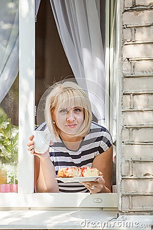 Young pretty blonde woman leaning through the window holding cup of tee and apple pie in hands smiling on sunny summer day Stock Photo