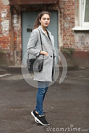 Lifestyle fashion portrait of beautiful young brunette woman in grey coat with black leather bag posing on street cloudy day Stock Photo