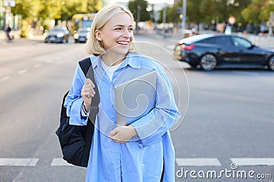Lifestyle portrait of candid young woman, walking on street, laughing and smiling, holding backpack and laptop, looking Stock Photo