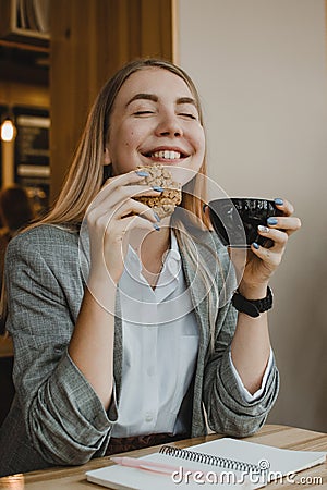 Lifestyle Portrait of smiling young casual blonde woman eating a cookie and drinking coffee, tea in cafe. Happy girl eating a diet Stock Photo