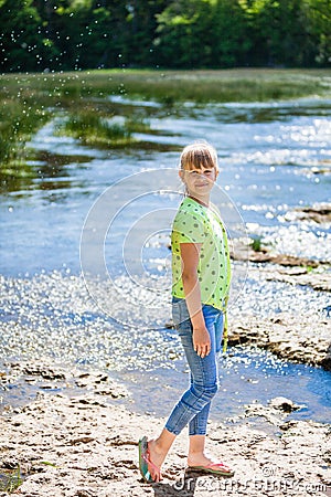 Portrait of little smiling girl child walking outdoor near calm lake at sunny day in spring. Teen is wearing jeans and green shirt Stock Photo