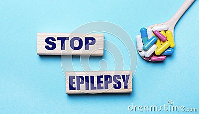 On a light blue background, bright multi-colored pills in a spoon and two wooden blocks with the text STOP EPILEPSY. Medical Stock Photo