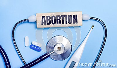 On a light gray background, a stethoscope, an electronic thermometer, pills, a wooden block with the text ABORTION. Medical Stock Photo