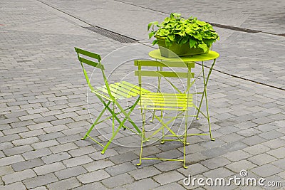 light green garden chairs and a folding table with a green flower box on the gray cobbled marketplace, art concept, color in Stock Photo