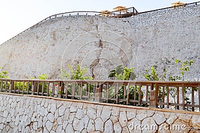 Light high stone wall with wooden rails and a sheer rock against the background of umbrellas from the sun and blue sky in a tropic Stock Photo