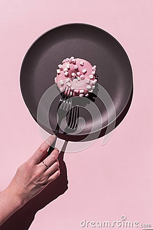 Light pink delicious donat with white marshmallows at the dark grey ceramic plate on the rosy background. Hand of woman Stock Photo