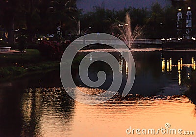 Light reflects fountain discharging water jet on to lake water surface in golden hour twilight. Beautiful Natural Landscape Stock Photo