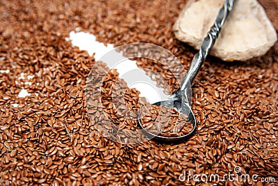 a light stone with a dark gray metal spoon on it lies against a background of brown flax seeds. On a spoon are flax seeds Stock Photo
