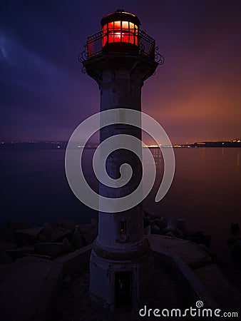 lighthouse on the seashore with red light at night against the background of the lights of the ships Stock Photo