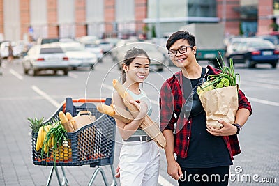 Likable asian couple hold paper eco bags with organic healthy food in hands while standing near store mall. Happy family Stock Photo