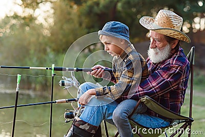 Likable respected 70-aged bearded grandfather holding on knees 10-aged cute grandson and teaching him to catch fish. Stock Photo