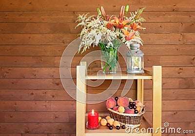 Lilies and astilbe flowers, burning lantern, candle and wicker basket with fruits stand on wooden shelves Stock Photo