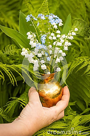 Lilies of the valley in May and forget-me-nots in a charming little jug held in a woman`s hand against the background of a garden Stock Photo