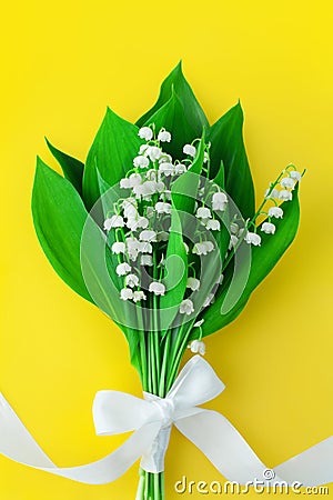 Lily of the valley flower bouquet & white bowknot on yellow background closeup, beautiful may lilies bunch & green leaves, ribbon Stock Photo
