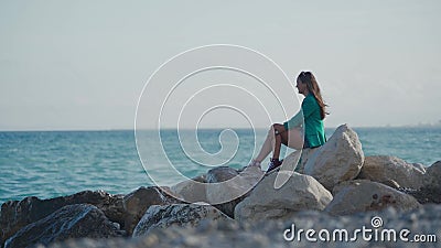 Linda mulher em tênis e uma blazer senta em uma praia rochosa e procurando ondas se espalhando contra as rochas Distante video estoque Linda mulher em tênis e uma blazer senta em uma praia rochosa e procurando ondas se espalhando contra as rochas Distante video estoque