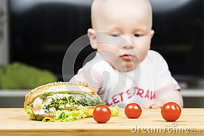 Litlle infant caucasian boy is trying to steal one unhealthy delicious hotdog with green salad from the table Stock Photo