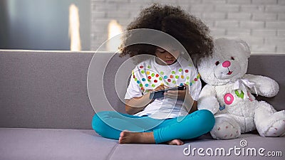 Little African female kid sitting on sofa playing on cellphone, gadget addiction Stock Photo