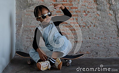 Little African kid boy wear cool gold sunglasses, necklace chain, jeans bib and sneaker smiling and sitting on skateboard Stock Photo
