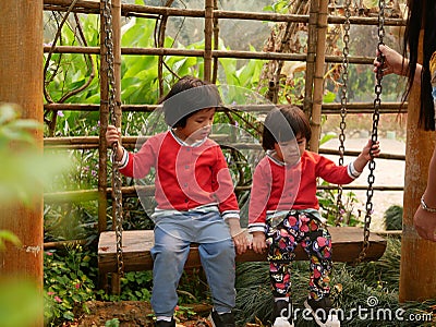 Little Asian baby girls, sisters, enjoy being in a swing together in a garden, with their mother watching close by Stock Photo