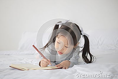 Little Asian girl lying on bed and writing alphabet on notebook Stock Photo