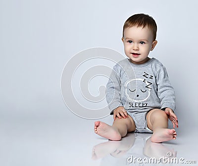 Little baby boy toddler in grey casual jumpsuit and barefoot sitting on floor and smiling over white wall background Stock Photo