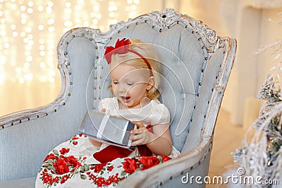 Little baby girl charming blonde in a red dress sitting in a chair against a background of Christmas trees and wonders present, i Stock Photo