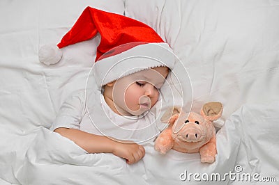 Little baby sleeping on white linen in the Santa hat with his toy pig, wich is the symbol of the year 2019. Stock Photo