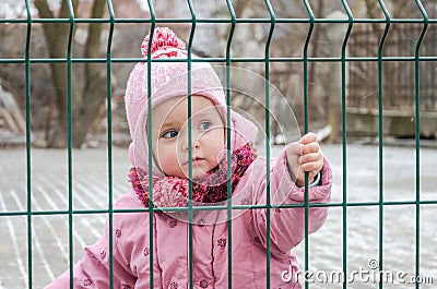 Little beautiful girl baby behind the fence, grid locked in a cap and a jacket with sad emotion on his face Stock Photo
