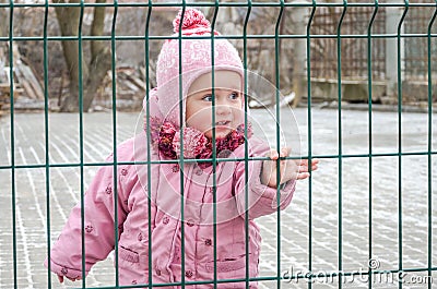 Little beautiful girl baby behind the fence, grid locked in a cap and a jacket with sad emotion on his face Stock Photo