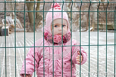 Little beautiful girl baby behind the fence, grid locked in a cap and a jacket with sad emotion on his face Stock Photo