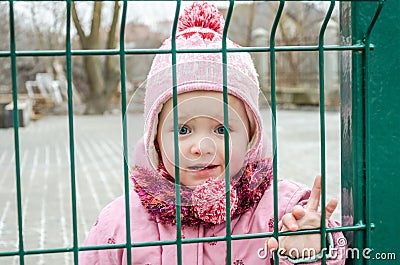 Little beautiful girl baby behind the fence, grid locked in a cap and a jacket with sad emotion on his face Stock Photo