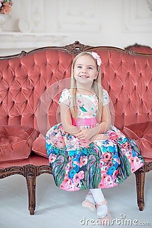 A little blond girl in colorful dress sitting on big sofa Stock Photo