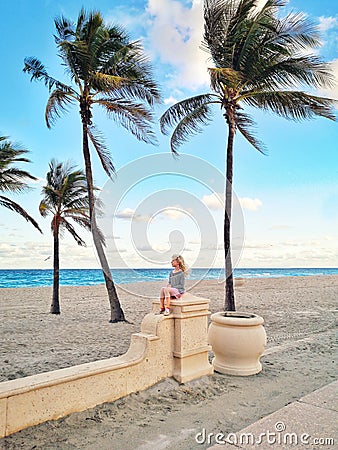 Little blonde Caucasian girl sitting relaxing dreaming on empty Hollywood ocean beach in Florida. Child among tall palm trees on Stock Photo