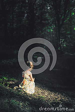 Little blonde girl in dress sits in sun light inside a dark creepy forest looking away to the trail. Girl alone in the woods Stock Photo