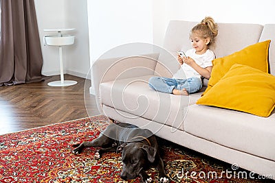 a little blonde girl at home on the sofa with a mobile phone and a big dog, children and gadgets Stock Photo