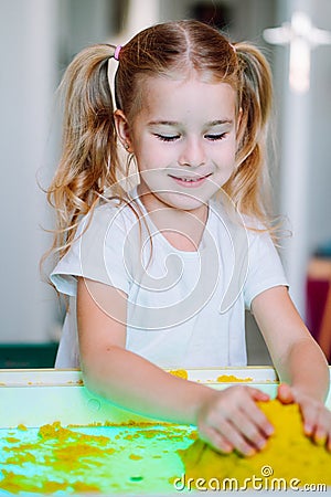 Little blonde girl play with yellow magic sand on white glowing table. Sensory development. Lessons in kindergarten Stock Photo