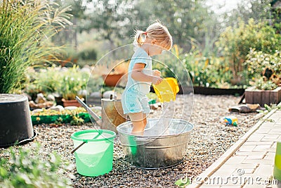 Little blonde girl playing at garden with water in a tin basin. Kids gardening. Summer outdoor water fun. Childhood in the country Stock Photo
