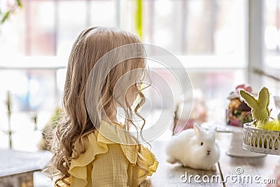 A little blonde girl in a yellow dress is sitting at a festive Easter table with rabbits.A baby and a rabbit. The concept of Stock Photo