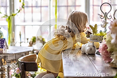 A little blonde girl in a yellow dress is sitting at a festive Easter table with rabbits.A baby and a rabbit. The concept of Stock Photo