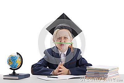 Little bored boy is balancing pencil on his nose while doing homework. Schoolboy in students hat at the table next to notebooks Stock Photo