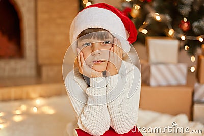 Little bored girl waiting for Christmas, sitting in festive living room, keeping hands on cheek, looks at camera with sad Stock Photo
