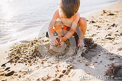 A little boy builds figures from the sand on the shore of the pond at sunset of the day, hands dig up the sand in crisp plan Stock Photo