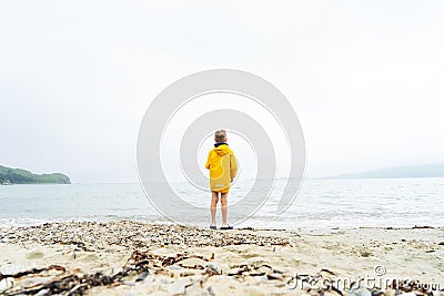 A little boy child in a yellow windbreaker stands on the sandy shore of the ocean all alone. Stock Photo