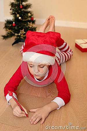 A little boy in Christmas clothes writes a letter to Santa lying on the floor Stock Photo
