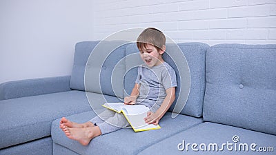 Little boy in gray clothes sitting on a gray sofa and learning to read a yellow book. Love for reading, family time. Self- Stock Photo