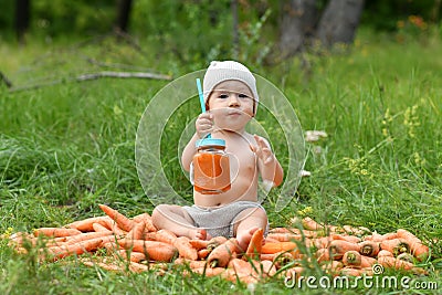 A little boy, one 1 year old, holding a jar of carrot juice on a pile of carrots in nature near a forest in a clearing and holds Stock Photo